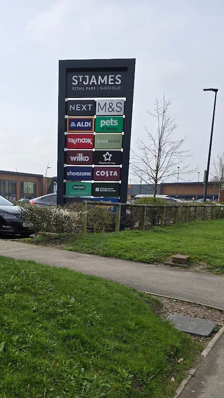 Sign for St. James Retail Park with store names like M&S, Aldi, and Costa, surrounded by greenery and parked cars.