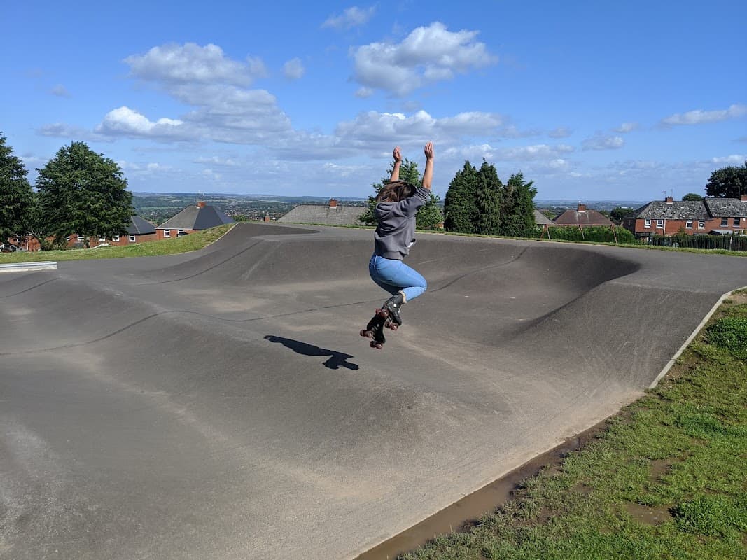 A person in roller skates jumps over a smooth, curved ramp in Grenoside Park, with houses and hills in the background.
