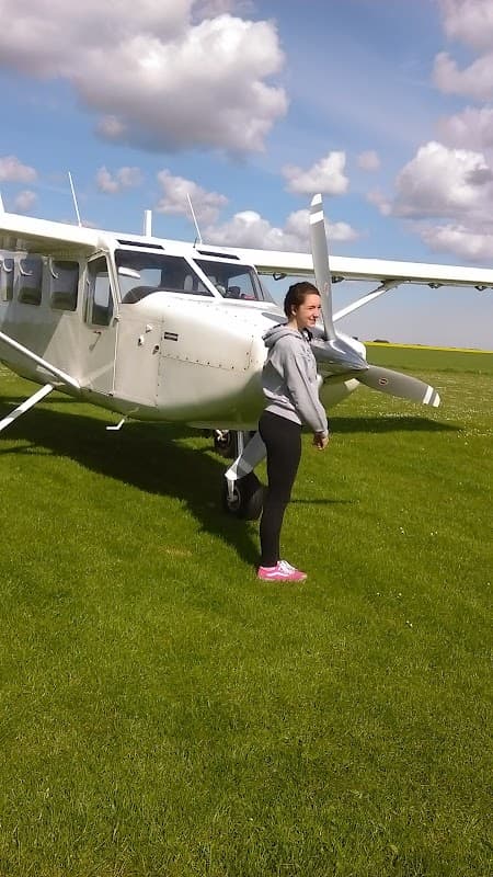 A woman stands beside a small white aircraft on a grassy field under a partly cloudy sky.