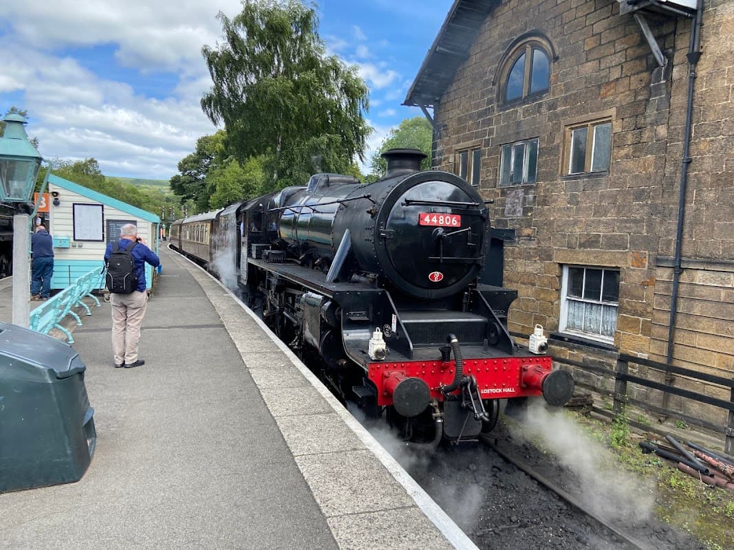 Steam train at Grosmont Station, with passengers waiting on the platform and historic stone buildings in the background.