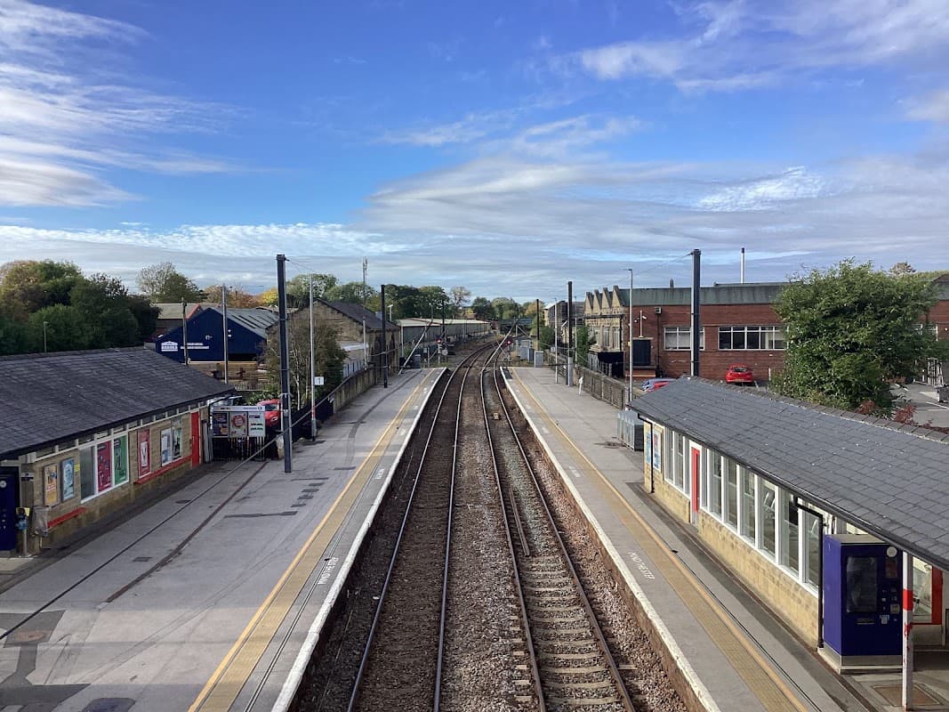 Railway tracks converge at Guiseley station, flanked by platforms and buildings under a blue sky with scattered clouds.