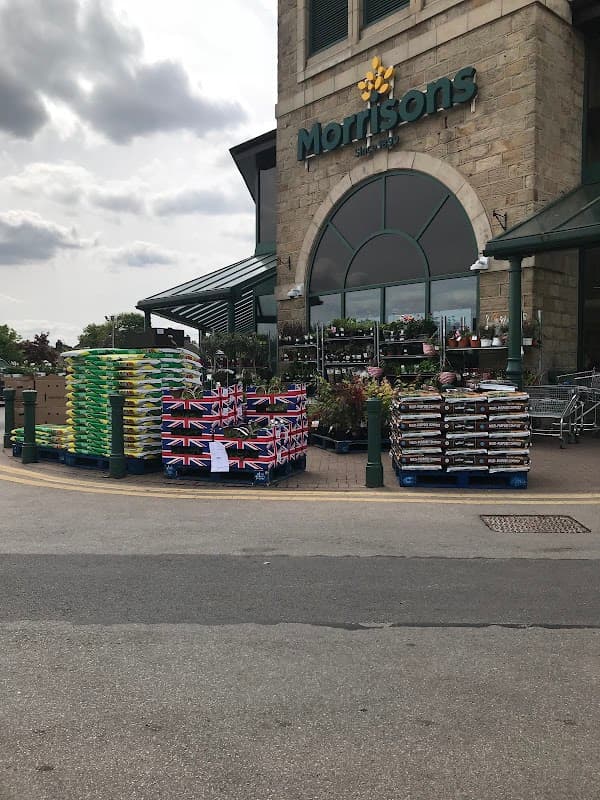 Morrisons store entrance with stacked pallets of plants, flags, and shopping carts in front under a cloudy sky.