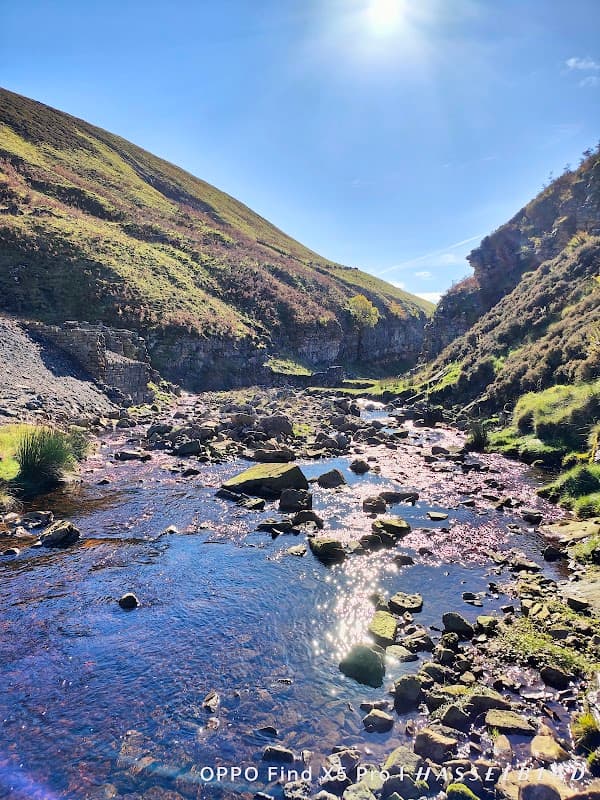 Sunlight glimmers on Gunnerside Beck, surrounded by lush green hills and rocky riverbanks in a serene landscape.