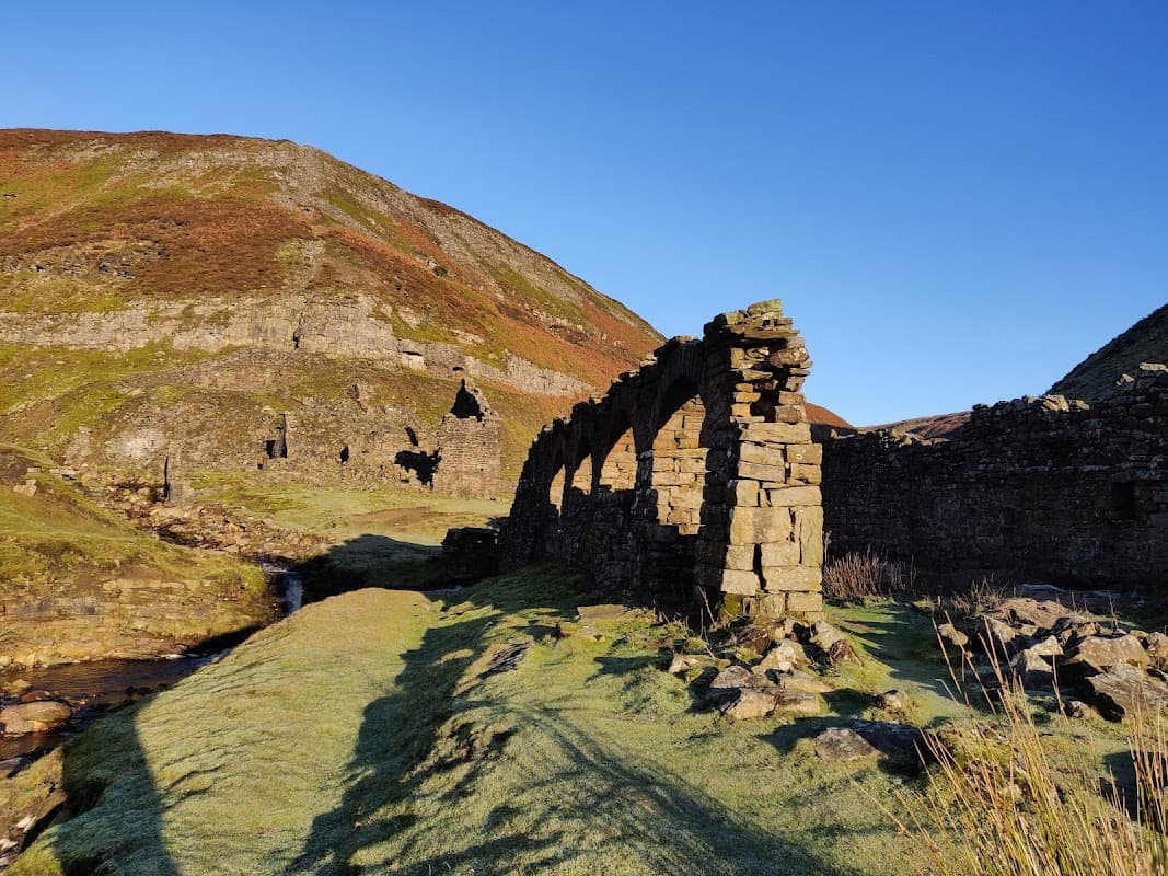 Stone ruins with archways beside a stream, set against a backdrop of green hills and clear blue sky.