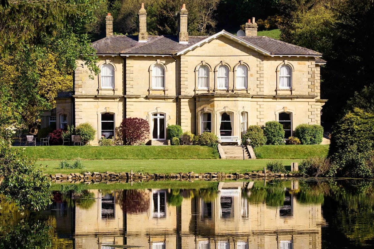 Elegant stone mansion with large windows, surrounded by lush greenery and reflecting in a calm lake.