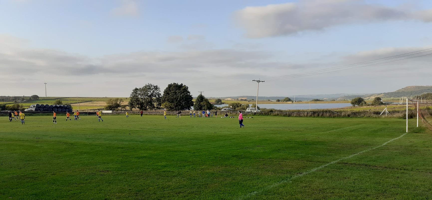 Green football pitch with players in action, surrounded by trees and a scenic landscape under a cloudy sky.