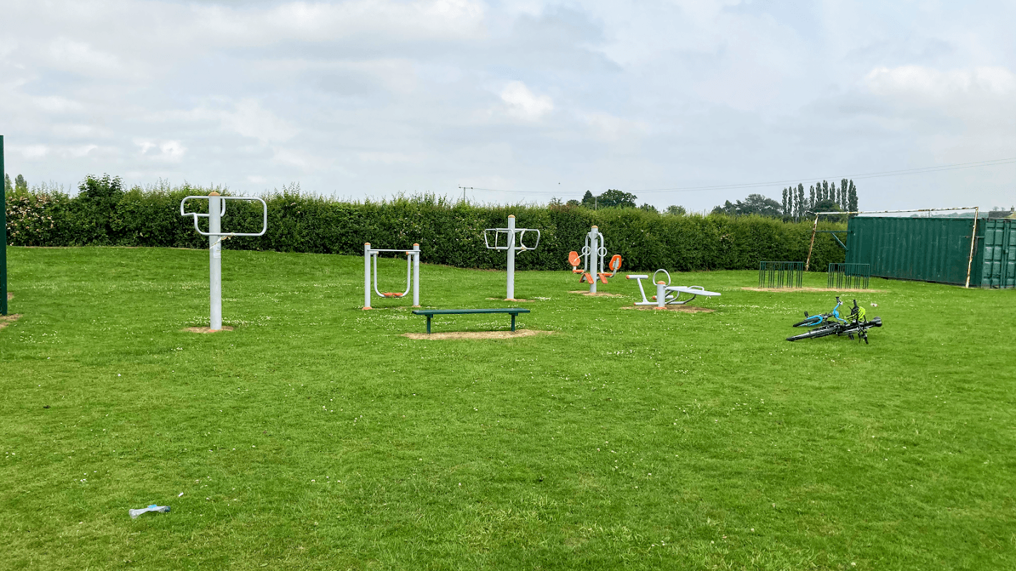 Outdoor fitness equipment on a grassy field, surrounded by hedges under a cloudy sky in Hambleton, Yorkshire.