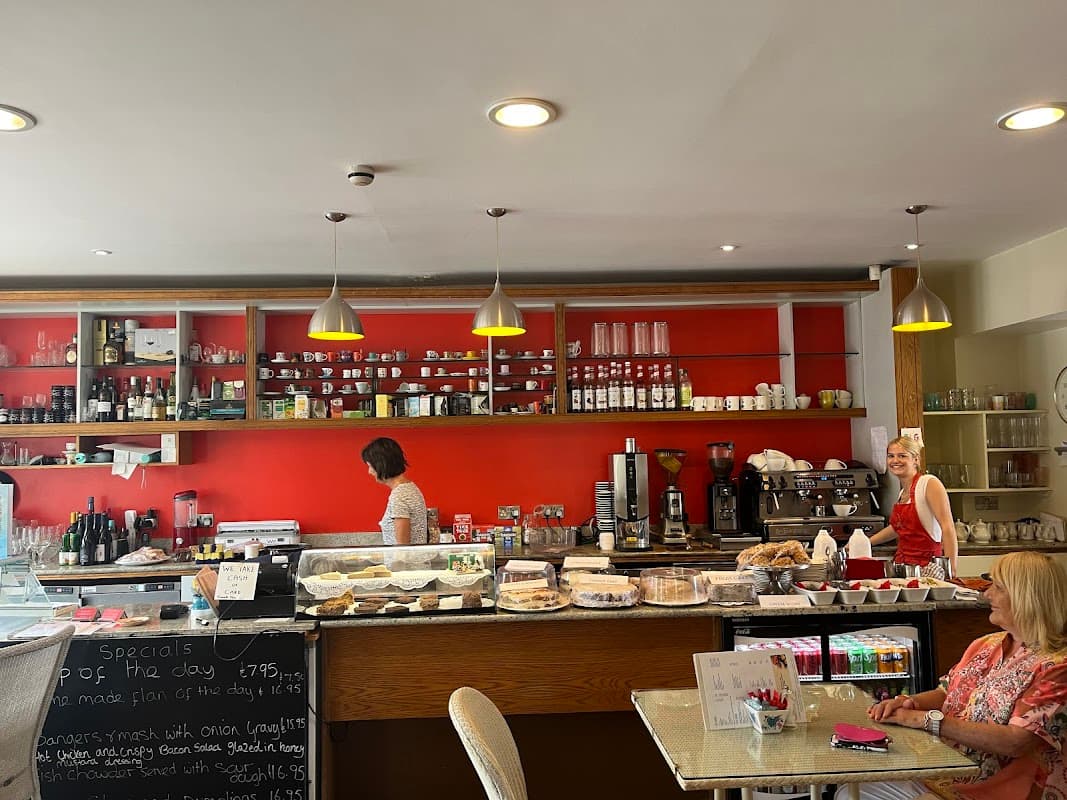 Bright coffee shop interior with a red wall, glass display of pastries, and two women chatting near the counter.