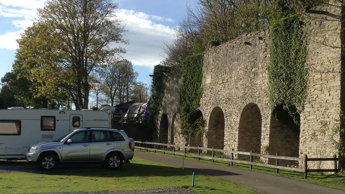 Caravans parked beside a stone wall with arches, surrounded by trees and a clear blue sky.