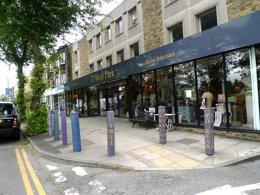 "Shopfront of 27 West Park Antiques & Home Interiors with bold signage, parking signs, and greenery lining the street."