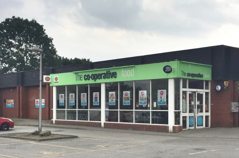 Co-op Food store with large green signage, glass entrance, and parking area, set against a cloudy sky.