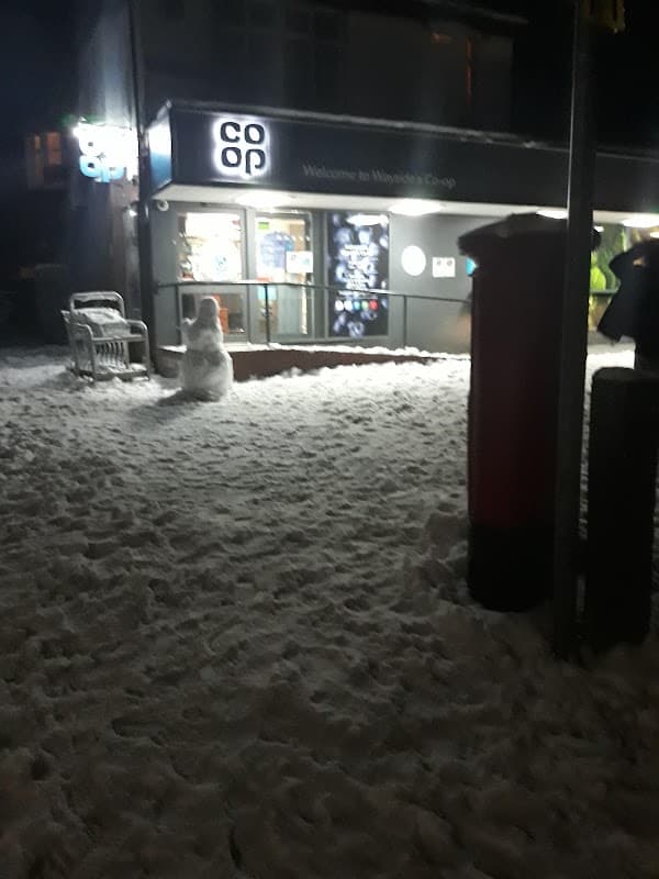 Co-op store entrance with snow-covered ground, a snowman, and a red post box at night.