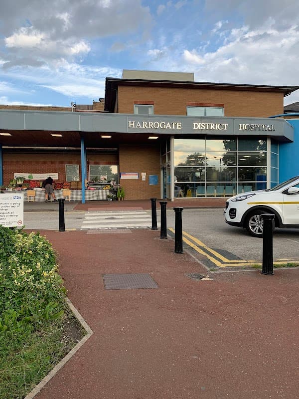 Harrogate District Hospital entrance with a blue facade, sign, and parked cars, surrounded by greenery.