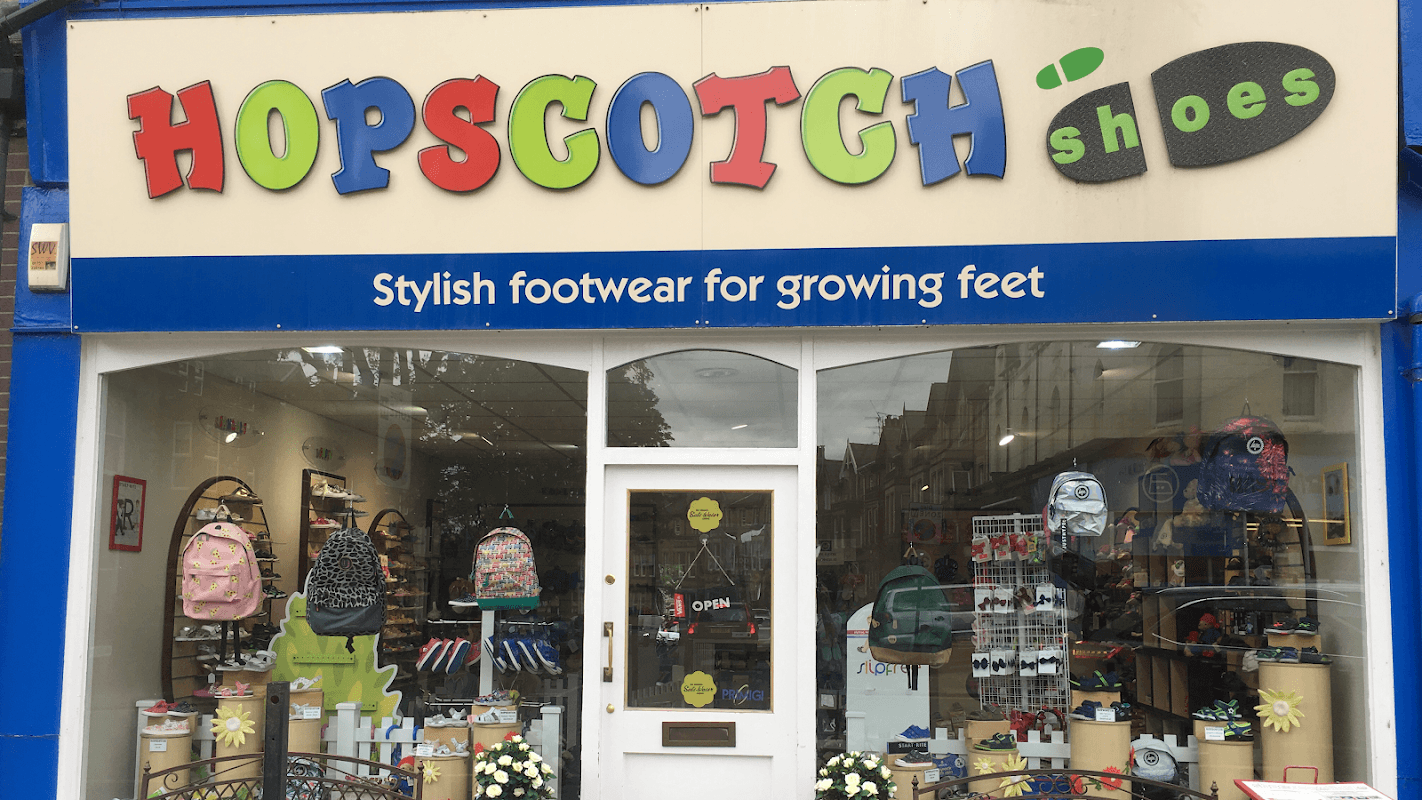Bright shopfront with "HOPSCOTCH shoes" sign, showcasing colorful footwear and backpacks in the window display.