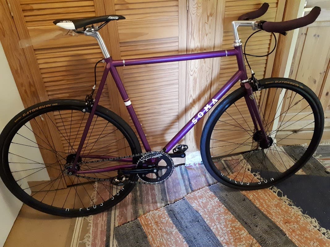 Purple fixed-gear bike with black wheels, leaning against a wooden wall, on a patterned rug.