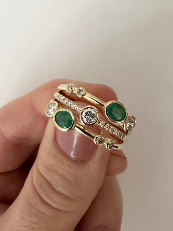 Gold rings featuring emeralds and diamonds, stacked together, held between fingers against a white background.