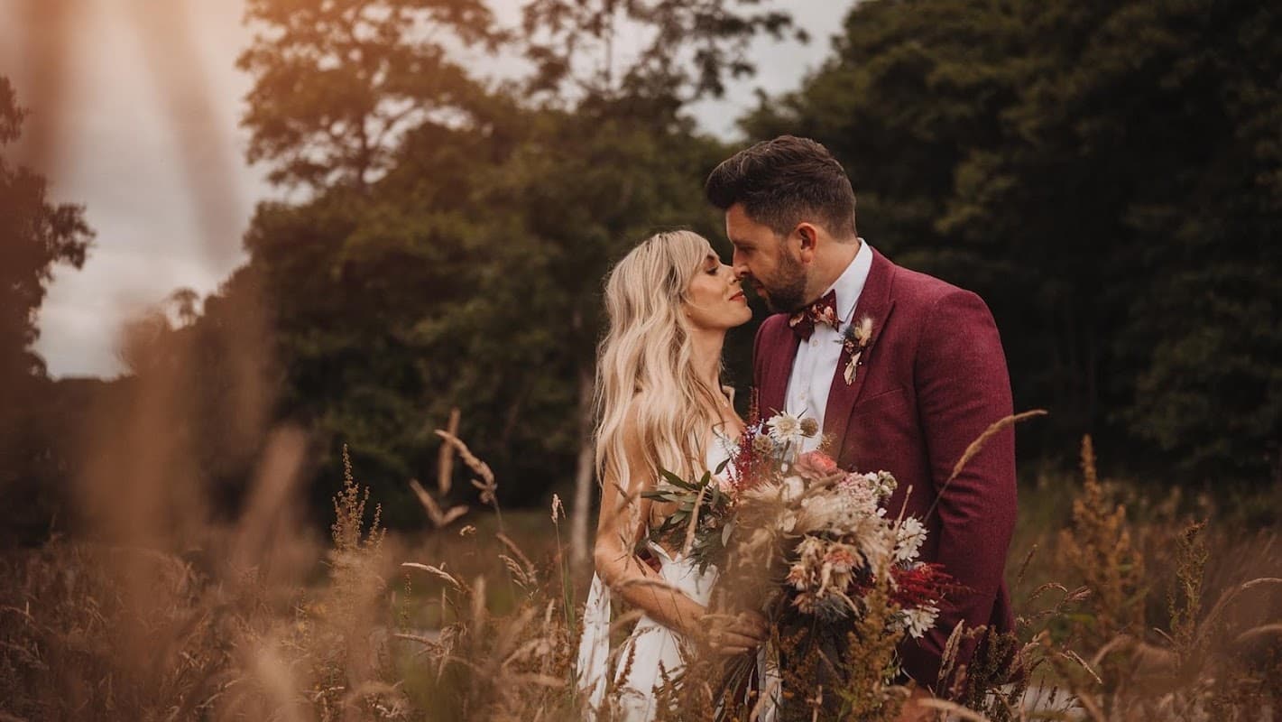A couple shares a kiss in a field, surrounded by tall grass and holding a vibrant bouquet.