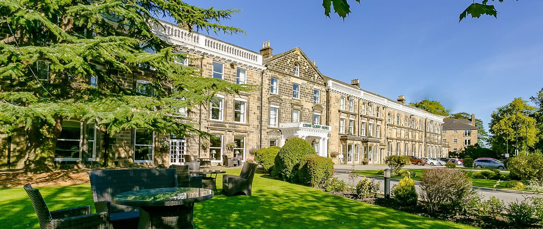Cedar Court Hotel exterior with manicured lawns, seating area, and clear blue sky in Harrogate, Yorkshire.