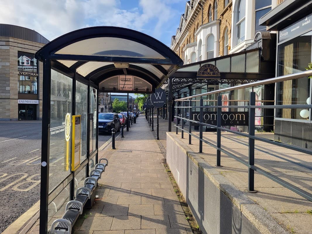 Bus stop shelter with seating, lined pavement, and shops in Harrogate, Yorkshire, under a partly cloudy sky.