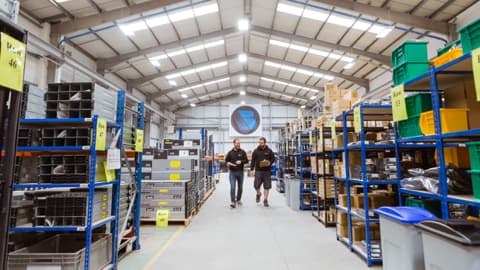 Two employees walk through a spacious warehouse filled with shelves of tech and electronics supplies.