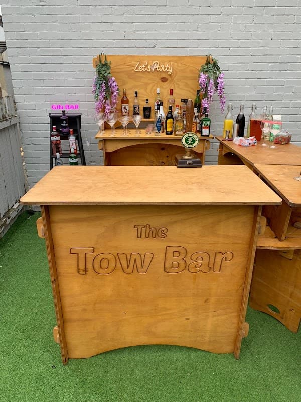 Wooden bar setup with "The Tow Bar" sign, shelves of drinks, and decorative flowers in a green outdoor space.