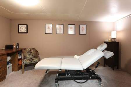 Treatment room with a white massage table, a desk, chair, and framed anatomical art on the walls. Soft lighting.