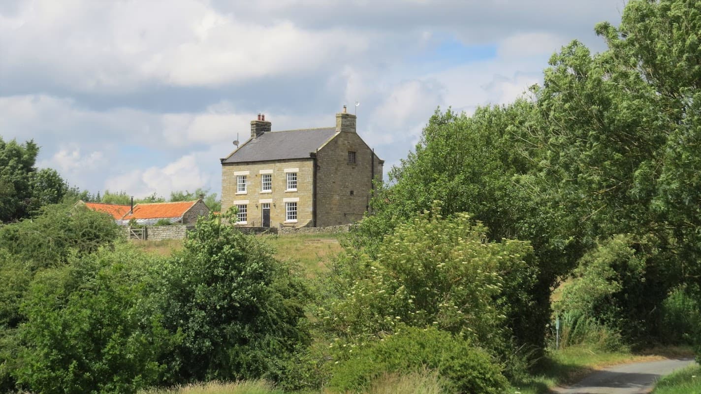Thirley Cotes Farm Cottages, a stone building surrounded by greenery and trees, with a distant red-roofed structure.