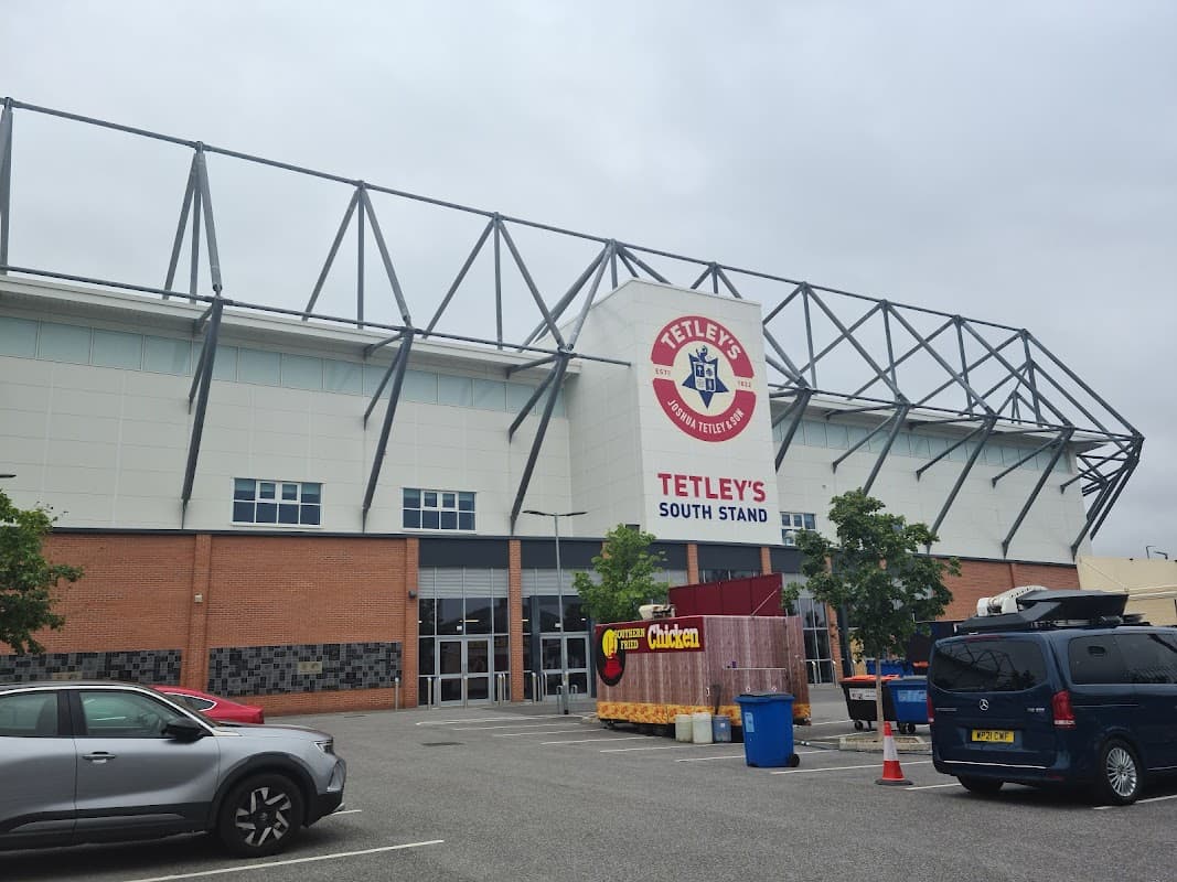 Tetley's South Stand building with a car park in front, featuring parked cars and nearby trees under a cloudy sky.