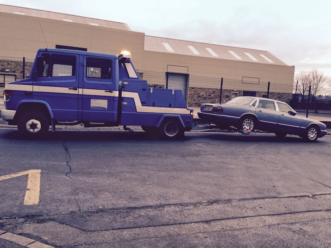 A blue tow truck pulling a silver car on a trailer in a parking lot, with a building in the background.