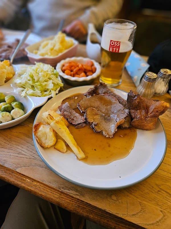 A plate of roast beef with gravy, Yorkshire pudding, potatoes, carrots, cabbage, and a pint of beer on a wooden table.
