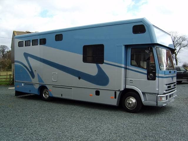 Blue and grey horsebox parked on gravel, with large windows and a streamlined design against a cloudy sky.