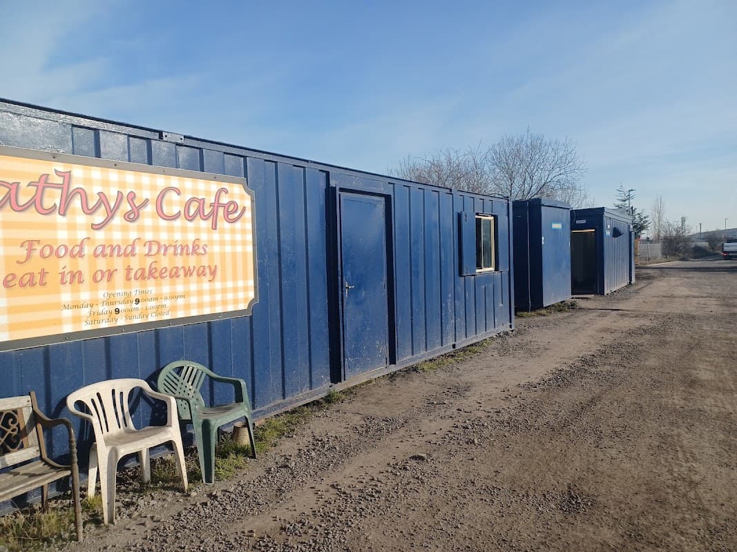 Blue shipping containers line a gravel path, with chairs in front of a cafe sign offering food and drinks.