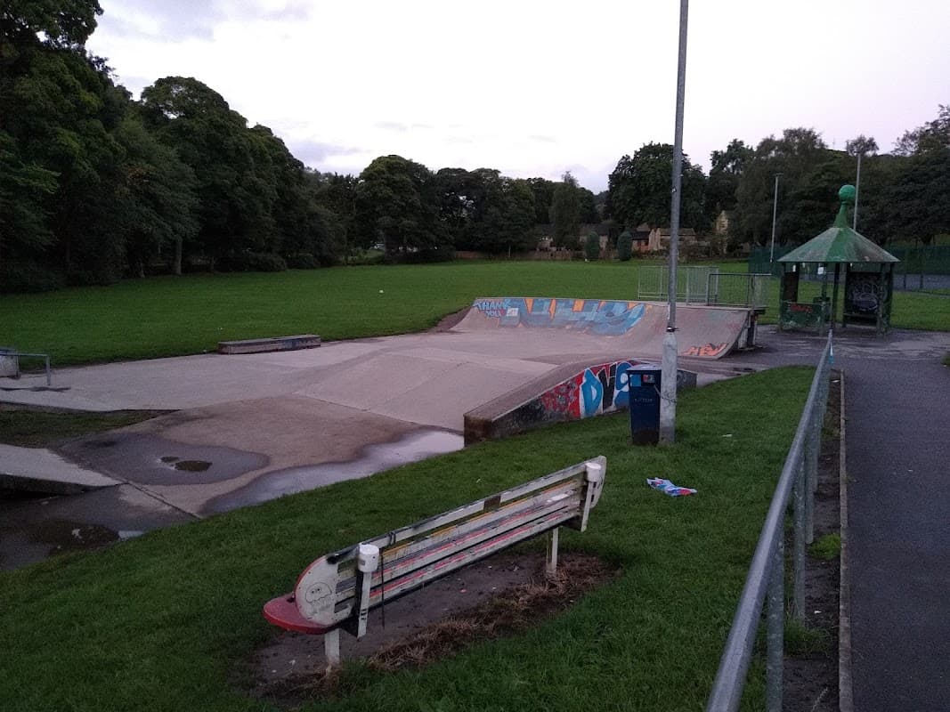 Skate park with a concrete ramp, colorful graffiti, green grass, and a nearby bench under a cloudy sky.
