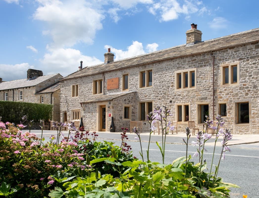 Stone building with large windows, surrounded by flowers and greenery, under a partly cloudy blue sky.