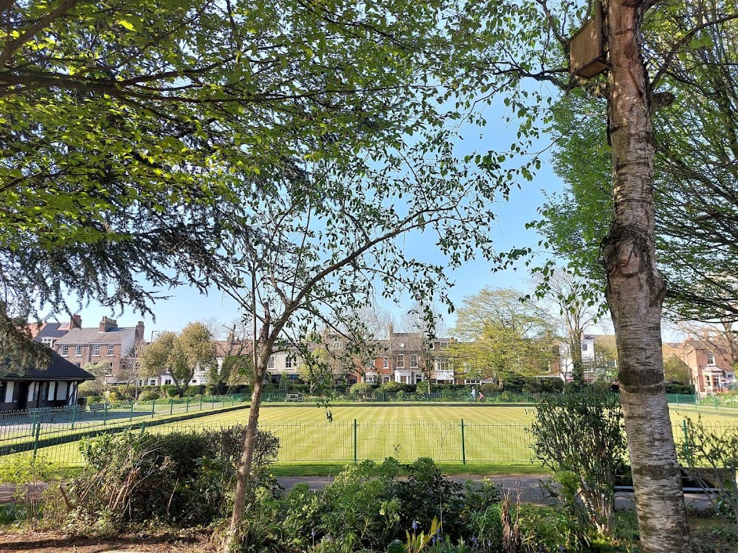 Lush green play area with trees, a manicured lawn, and residential buildings in the background under a clear blue sky.