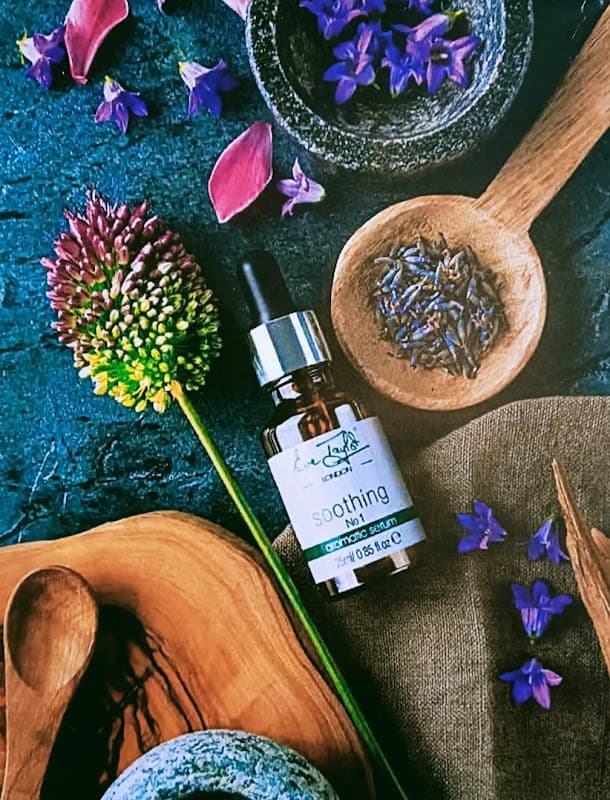 A soothing serum bottle surrounded by flowers, herbs, and wooden bowls on a dark textured surface.