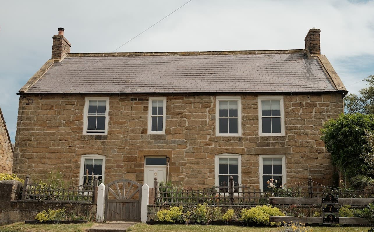 Stone cottage with a slate roof, white windows, and a wooden gate, surrounded by gardens in a rural setting.