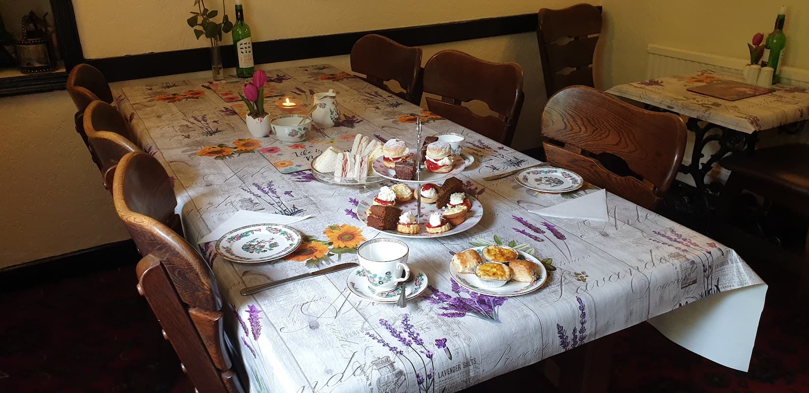 A dining table set with cakes, sandwiches, and tea, surrounded by wooden chairs in a cozy bar setting.