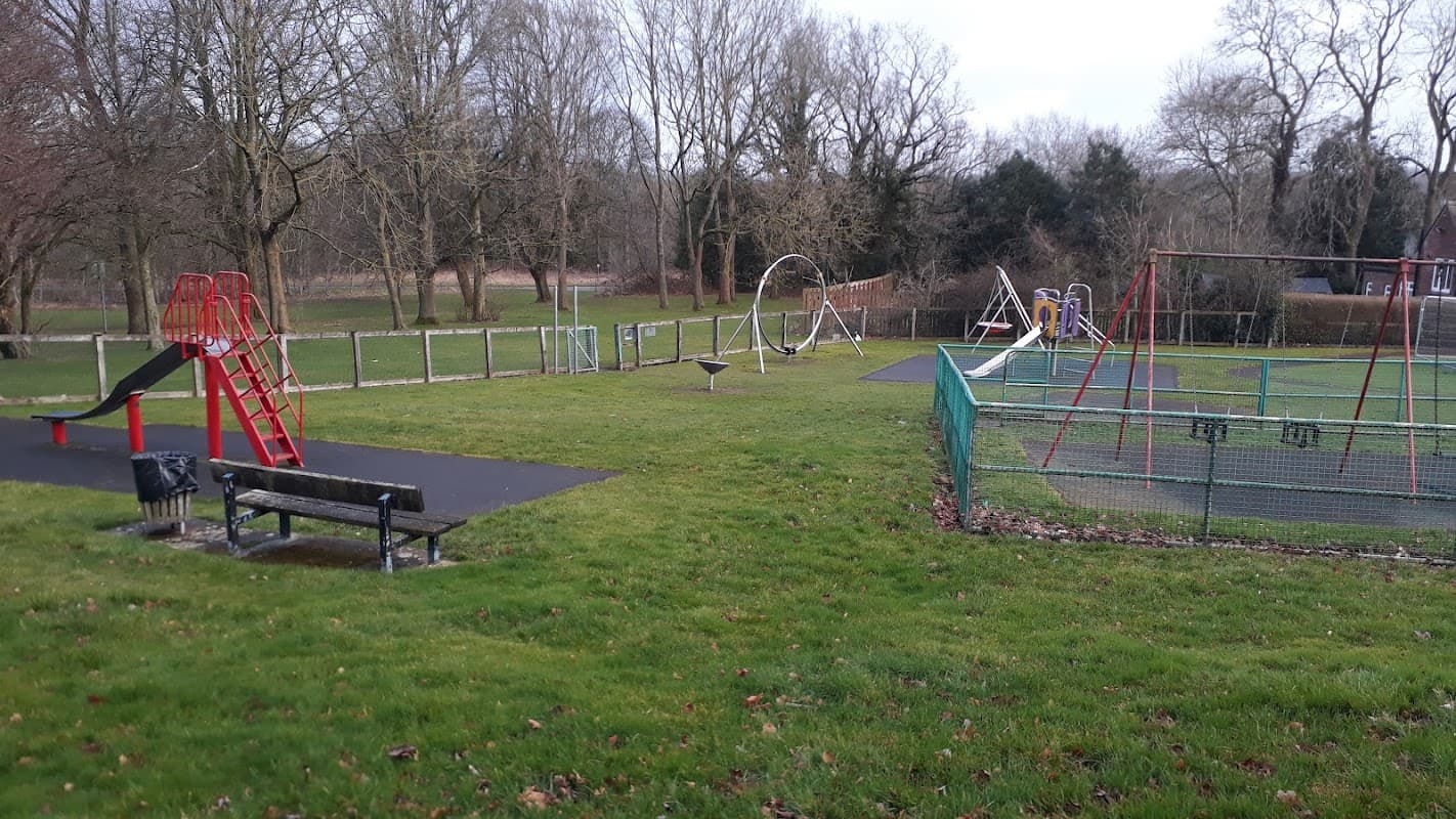 Playground with climbing frame, swings, benches, and open grassy areas surrounded by trees in Hipswell, Yorkshire.