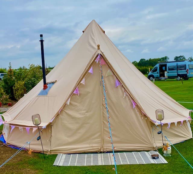 A beige bell tent adorned with bunting, set on green grass with a van parked nearby under a cloudy sky.