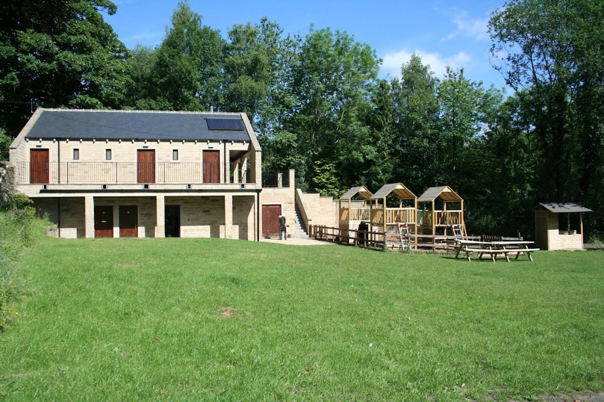 Modern building with wooden doors, surrounded by green grass and trees, featuring picnic tables and wooden structures nearby.