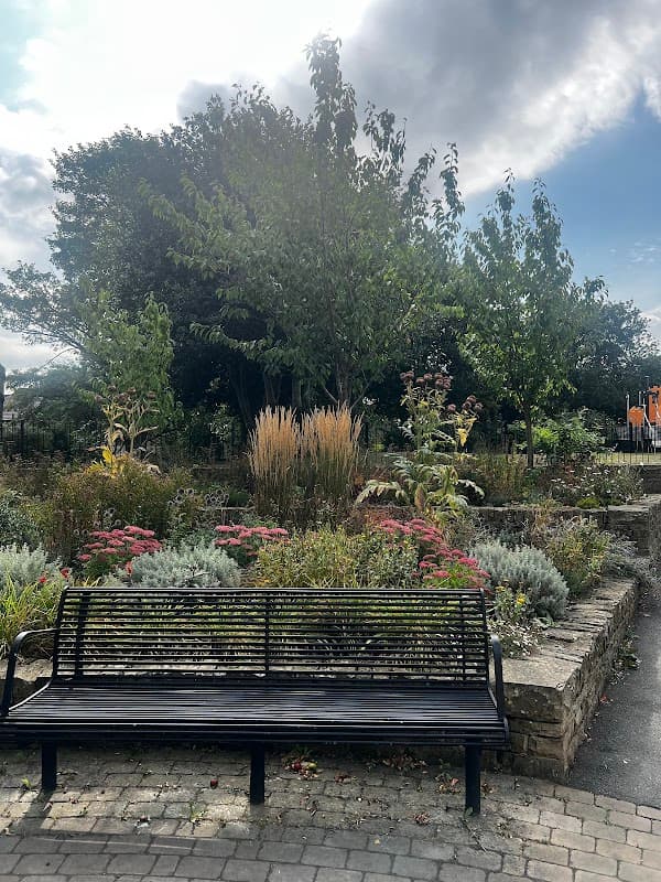 Park scene featuring a black bench surrounded by colorful flowers, shrubs, and lush greenery under a cloudy sky.