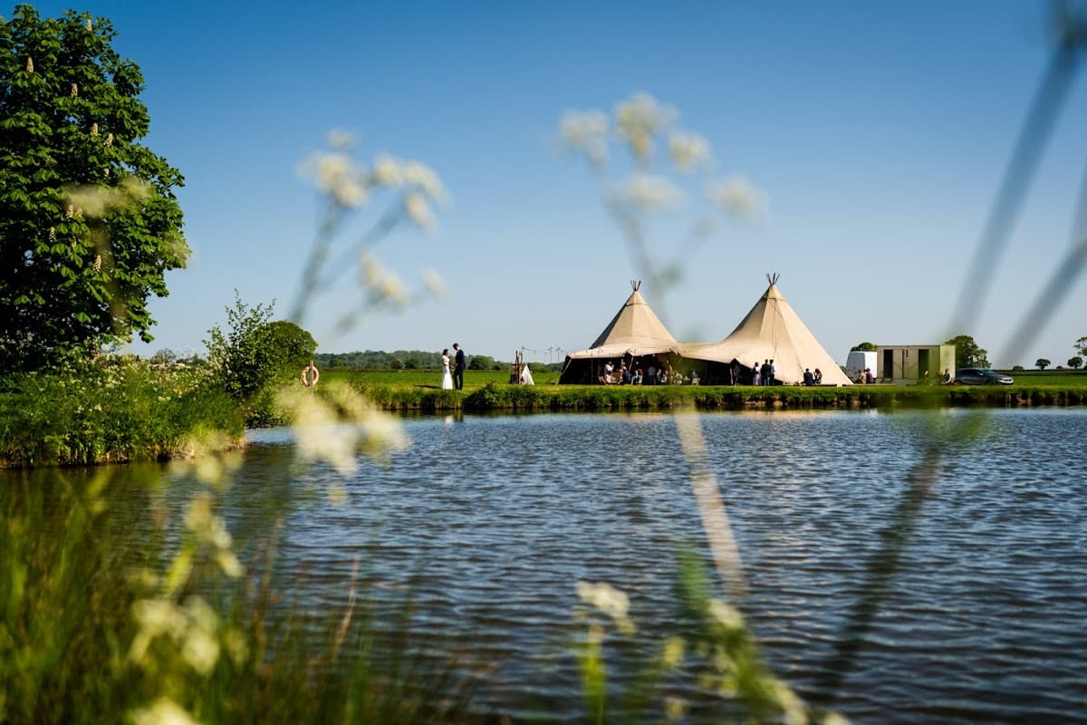 Tipi tents by a serene lake, surrounded by lush greenery and clear blue skies, perfect for a wedding venue.