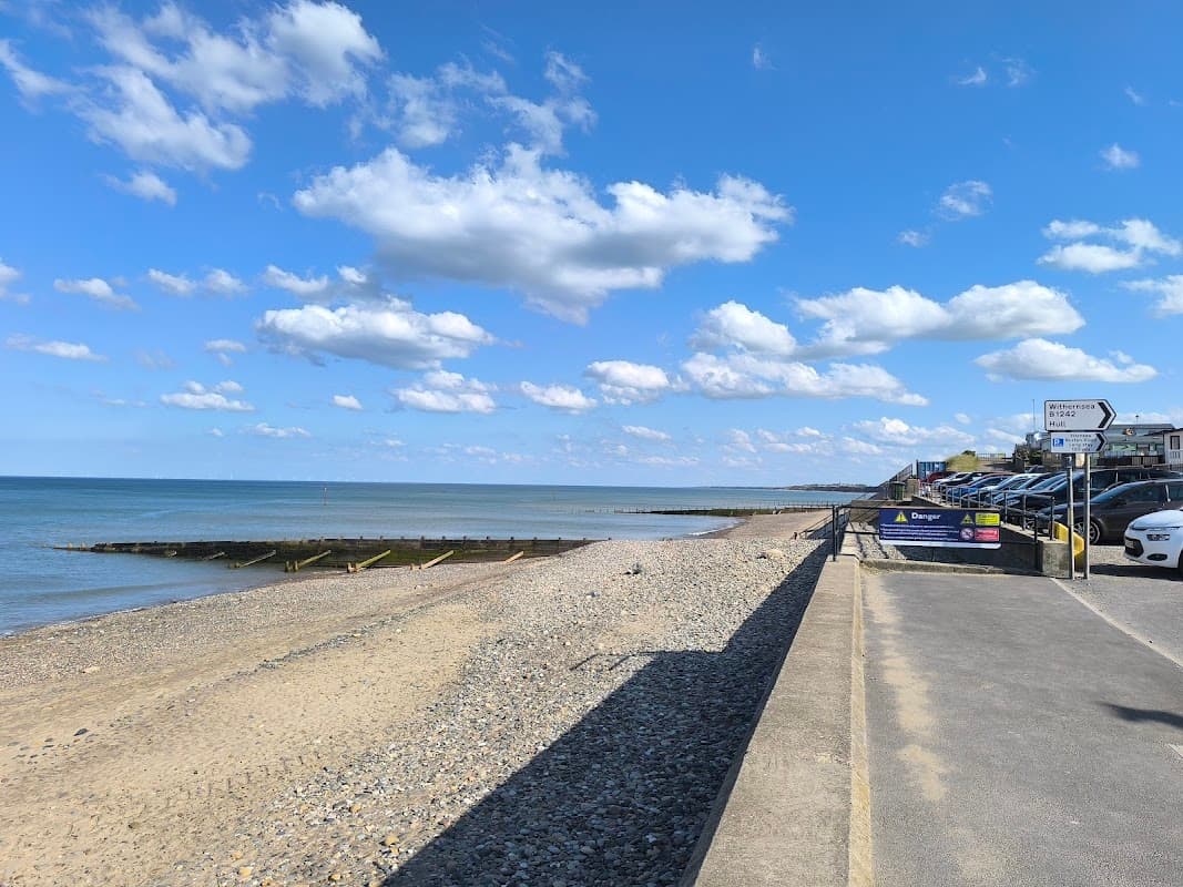 Coastal view with a sandy beach, pebbles, calm sea, and a car park lined with vehicles under a blue sky.