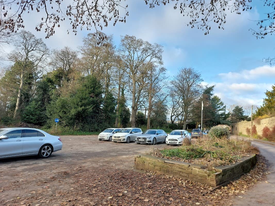 Several parked cars in a gravel lot surrounded by trees and a stone wall under a partly cloudy sky.