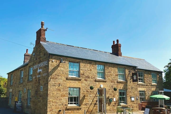 Historic stone inn with green-framed windows, brick chimneys, and outdoor seating under a clear blue sky.