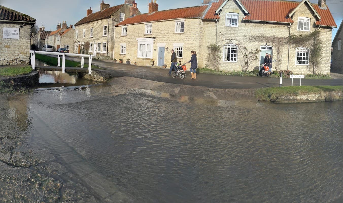 Charming stone cottages in Hovingham with a shallow stream, pedestrians, and strollers near a quaint village setting.