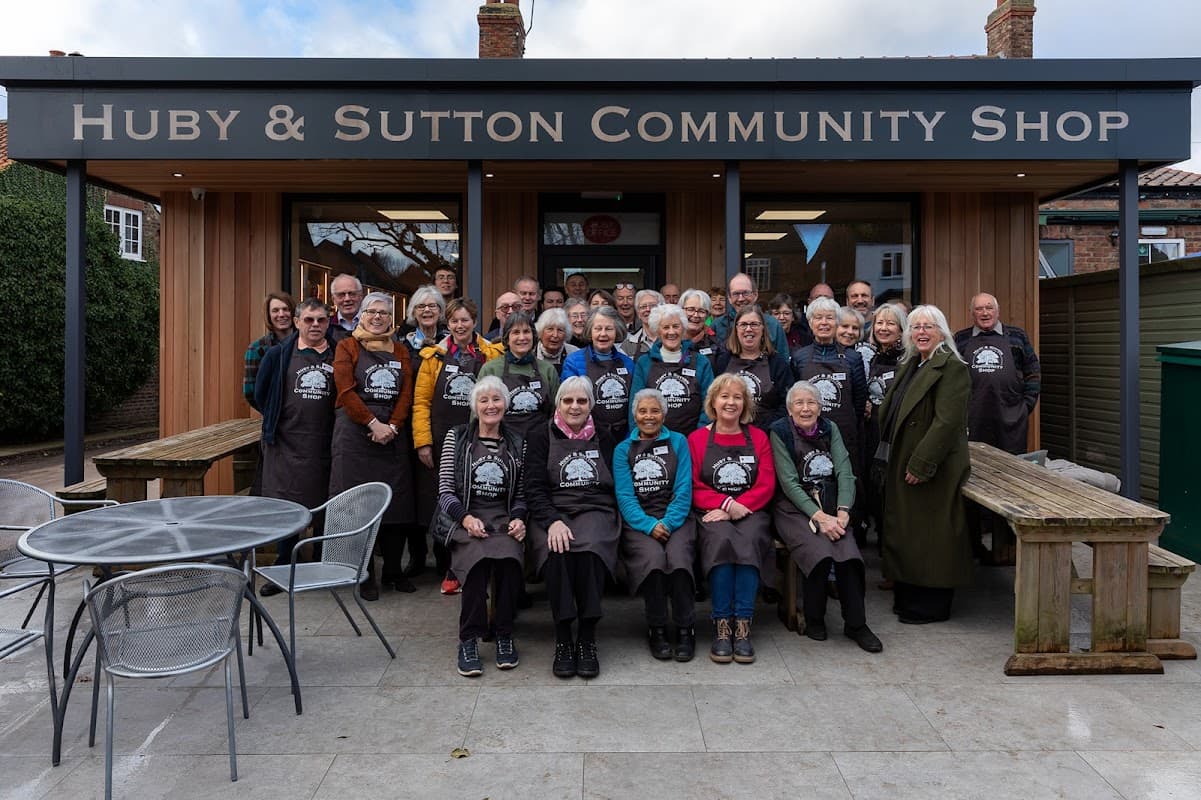 A large group of smiling volunteers in aprons stands outside the Huby & Sutton Community Shop, with tables and chairs nearby.