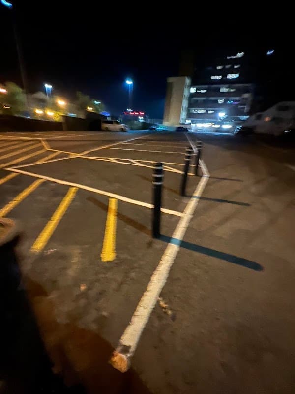Dark car park with yellow lines, black bollards, and illuminated buildings in the background. Nighttime setting.