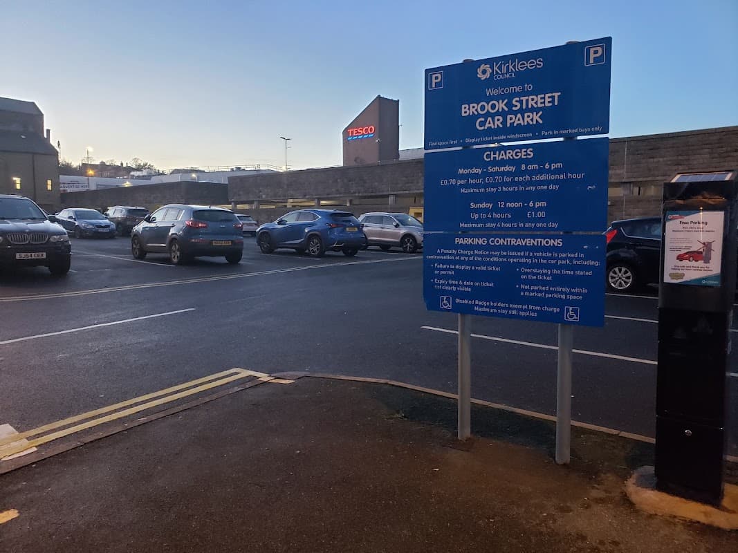 Brook Street Car Park sign with parking charges, surrounded by parked cars and a Tesco store in the background.